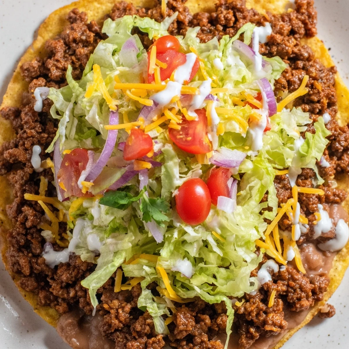 A close-up of a delicious homemade Beef Tostada with refried beans and fresh lettuce. Finished with a dollop of sour cream and a squeeze of lime for brightness.