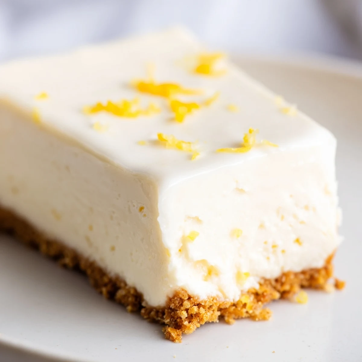 A slice of Rich Greek Yogurt Cheesecake served on a dessert plate, topped with a dollop of sweetened sour cream and lemon zest.